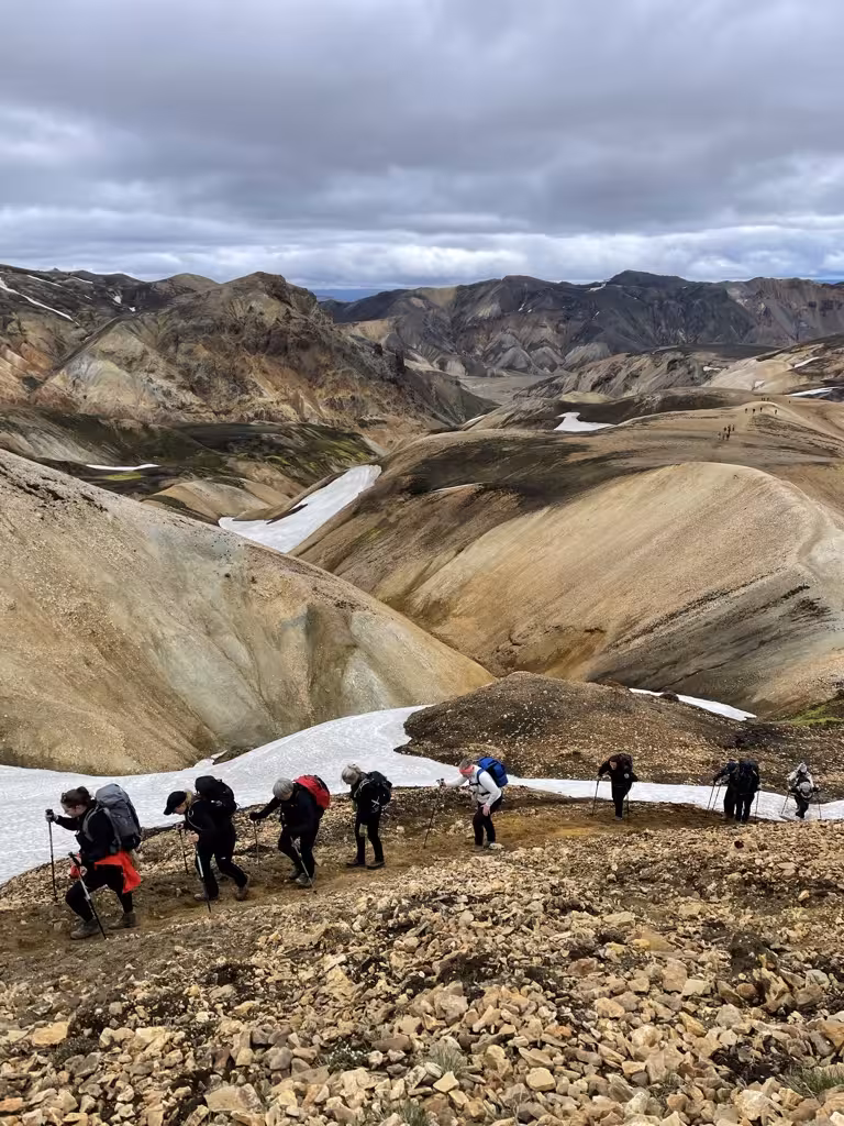 Women hikers ascend rocky terrain on the scenic Laugavegur trail, surrounded by Iceland's rugged mountains and snow patches.