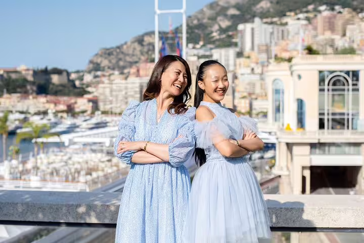Two women in light blue dresses standing back-to-back, smiling by the scenic waterfront in Nice with cityscape views.