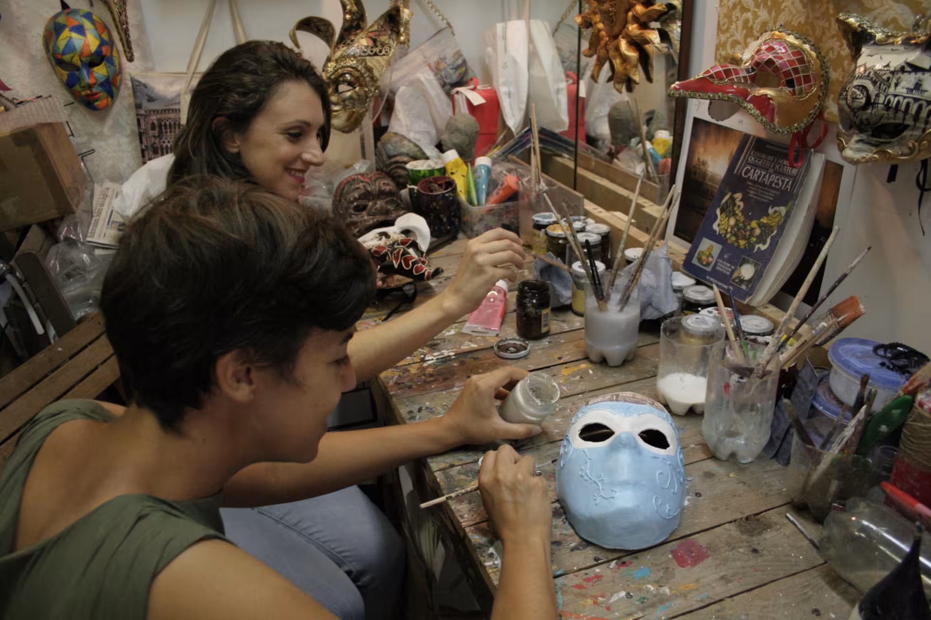 Two women painting masks with detailed designs in an immersive artisan mask decoration workshop environment.