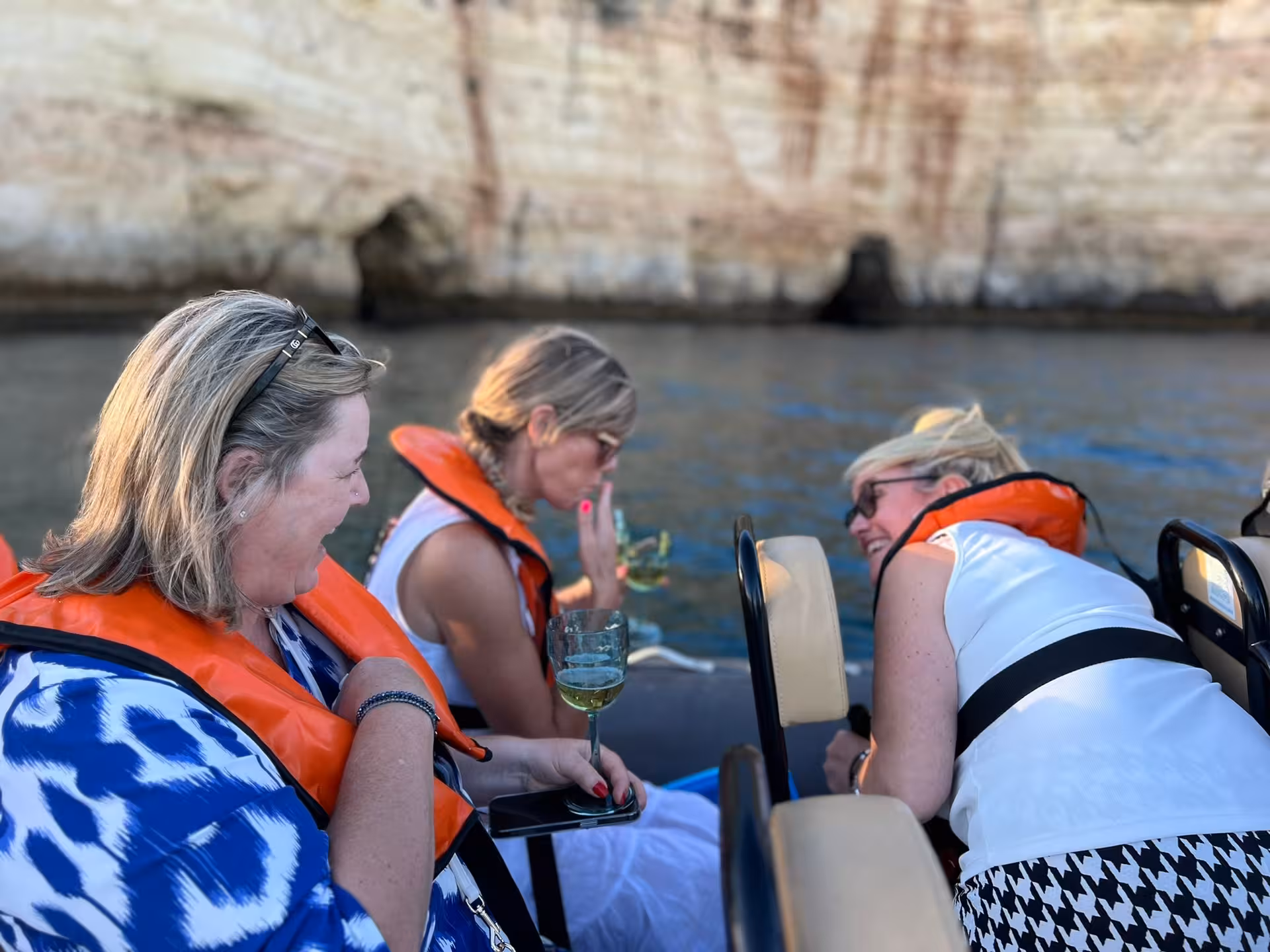 Women wearing life jackets laugh and enjoy wine on a private Algarve boat charter beneath dramatic coastal cliffs