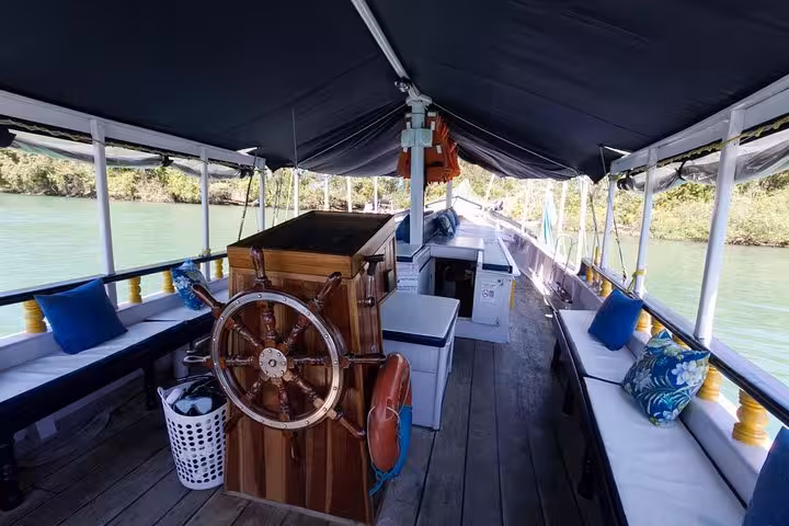 Interior view of a wooden boat with a steering wheel, cushioned seating, and scenic river surroundings for a relaxing tour.