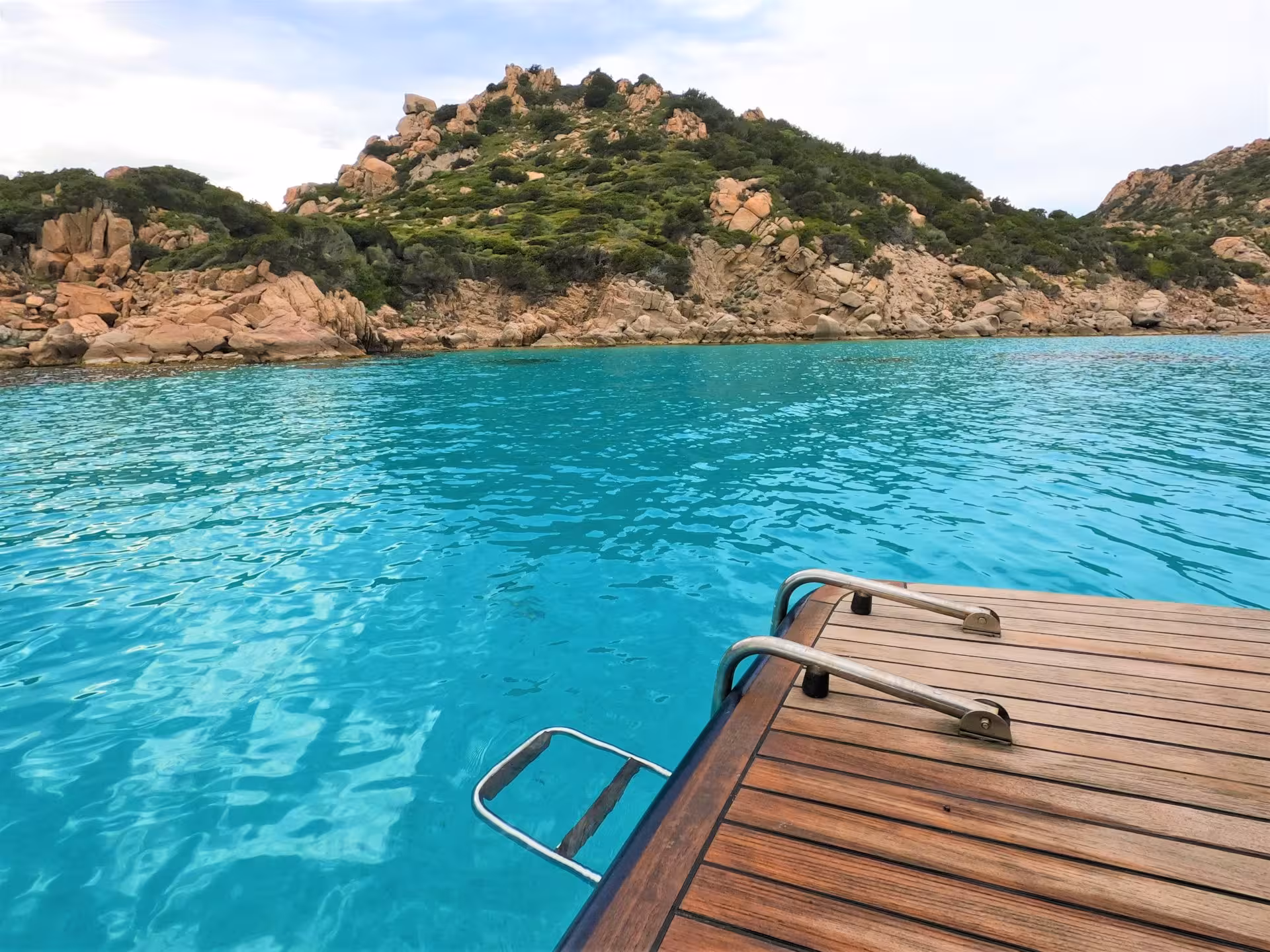 Wooden boat platform with ladder leading into clear blue waters, surrounded by rugged hills in La Maddalena Archipelago.