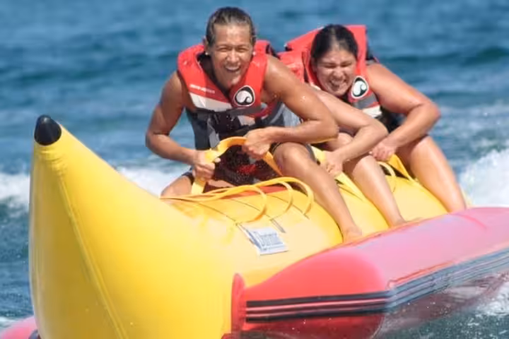 Two excited friends in life jackets ride a yellow banana boat across clear blue sea on a thrilling water sports adventure tour