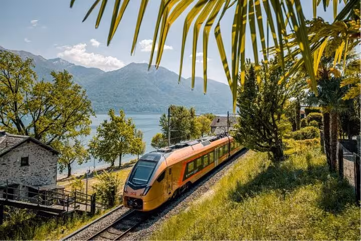 Scenic train journey along Lake Maggiore with a yellow train passing through lush greenery and mountain backdrop.