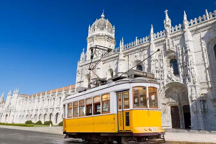 Historic yellow tram passing in front of the ornate Jerónimos Monastery in Belém, Lisbon, under a clear blue sky.