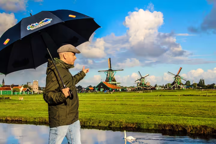 Man with umbrella pointing at iconic Zaanse Schans windmills under a bright sky, showcasing Dutch countryside charm.