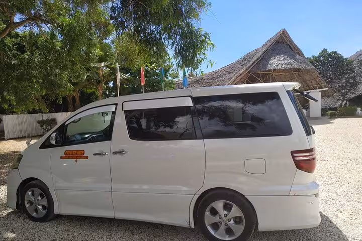 White minivan parked under tropical trees near traditional thatched building, providing 24/7 airport transfers to Nungwi hotels.
