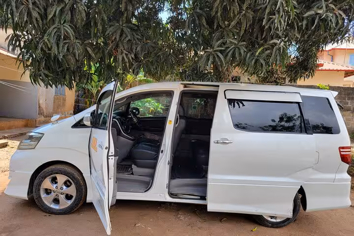 Open-door view of a white transfer van under trees, showcasing its interior for Zanzibar Island Shuttles.