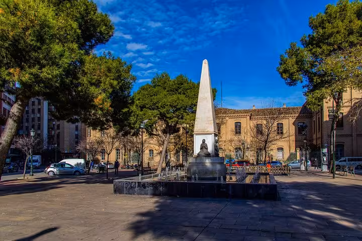 Sunny Zaragoza plaza with an obelisk monument, surrounded by lush trees and historic buildings on a city walking tour.
