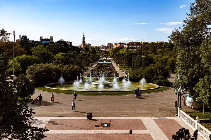 A panoramic view of Zaragoza's Parque Grande with its iconic fountain, lush greenery, and distant city skyline on a sunny day.