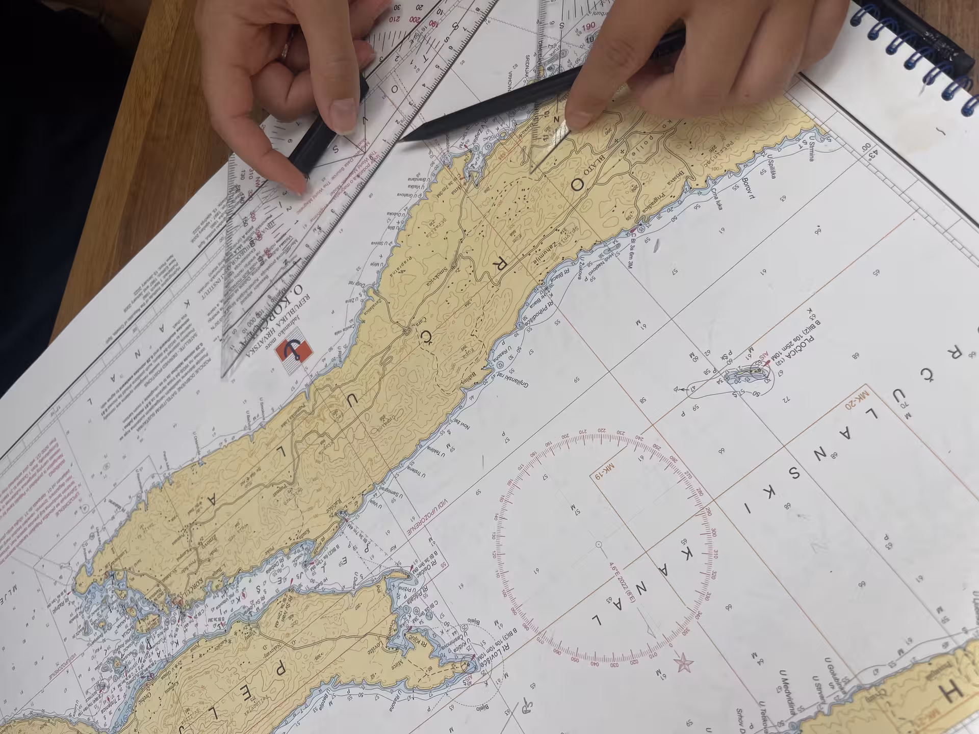 Hands plotting route on nautical chart with ruler and pencil, “Zero to hero” sailing course navigation lesson