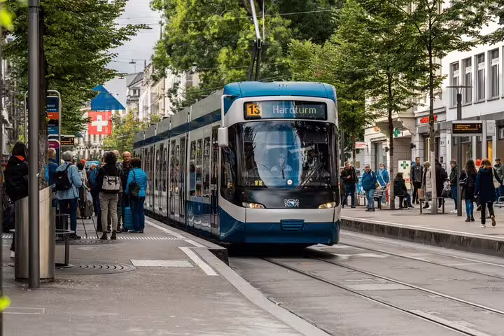 Blue tram navigating through a bustling Zurich street with people and Swiss flag in view.