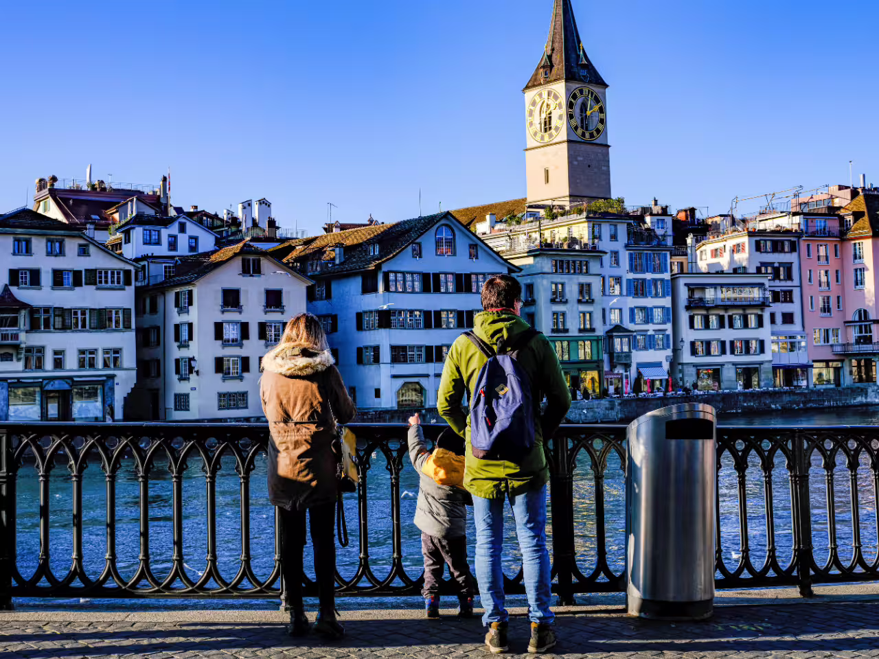 Family admires scenic Zürich riverfront and St. Peter's Church during a private half-day city tour.