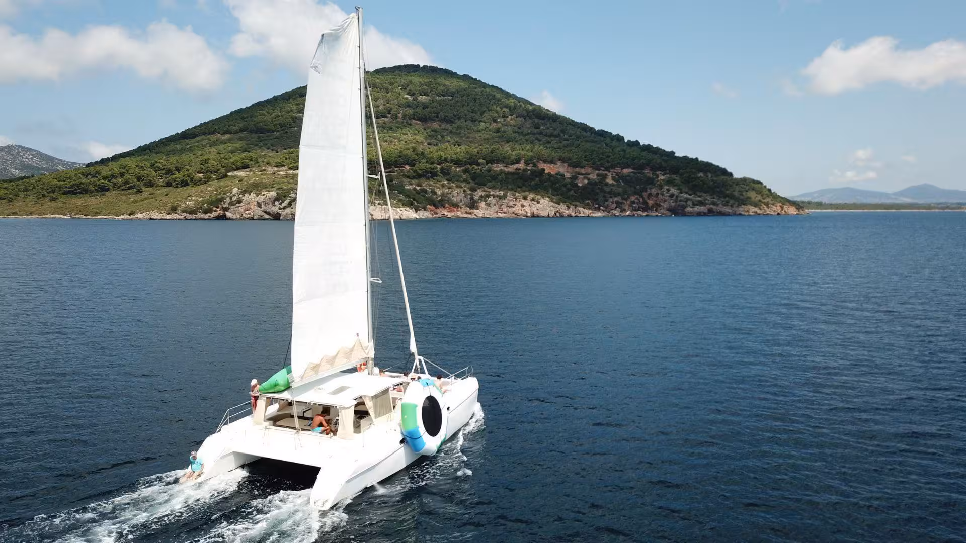Sailing catamaran gliding through calm blue waters with lush green hills in Porto Conte Park, Alghero tour.