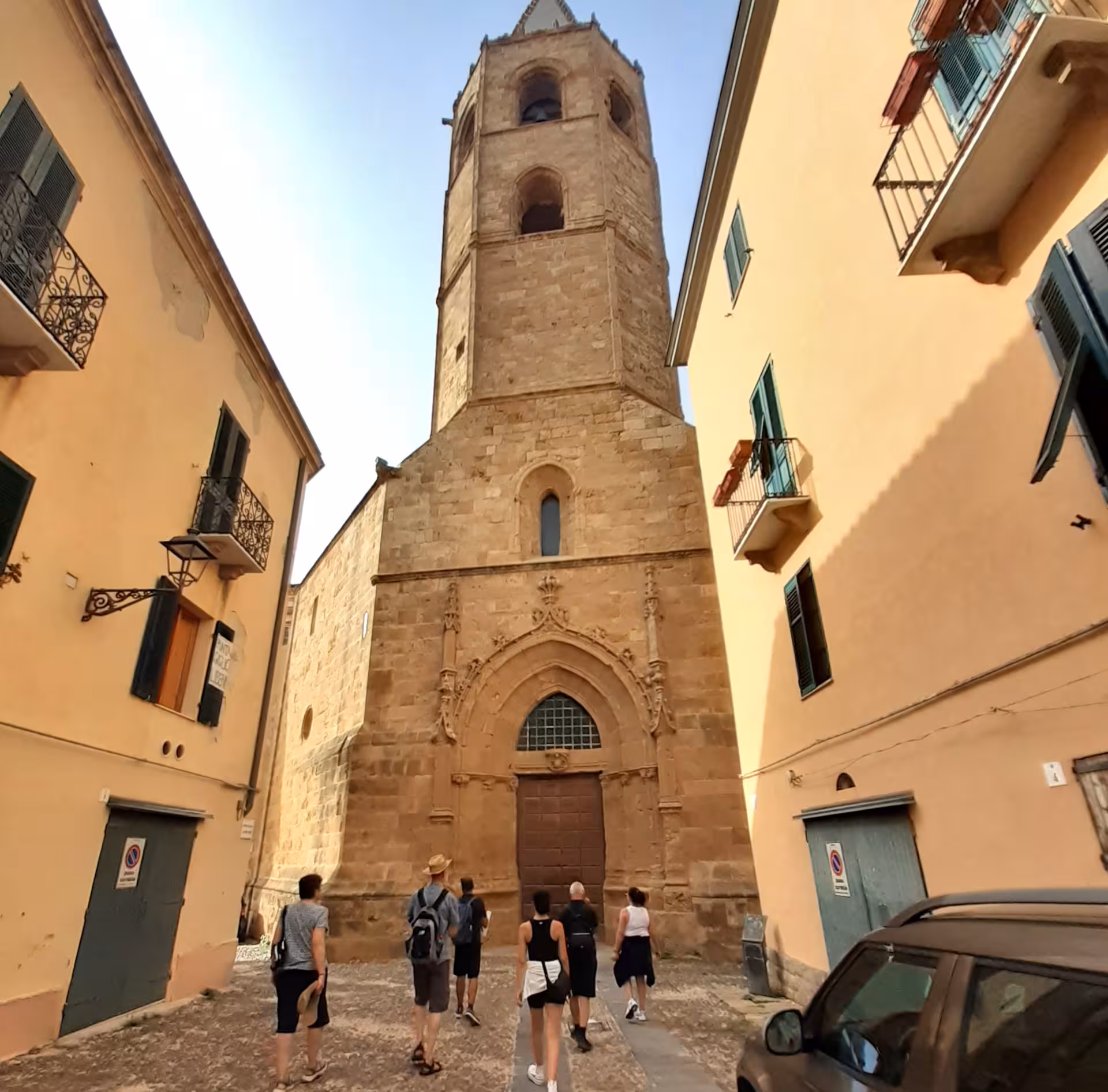 Tourists explore Alghero's historic center, admiring the towering architecture of a medieval stone church.