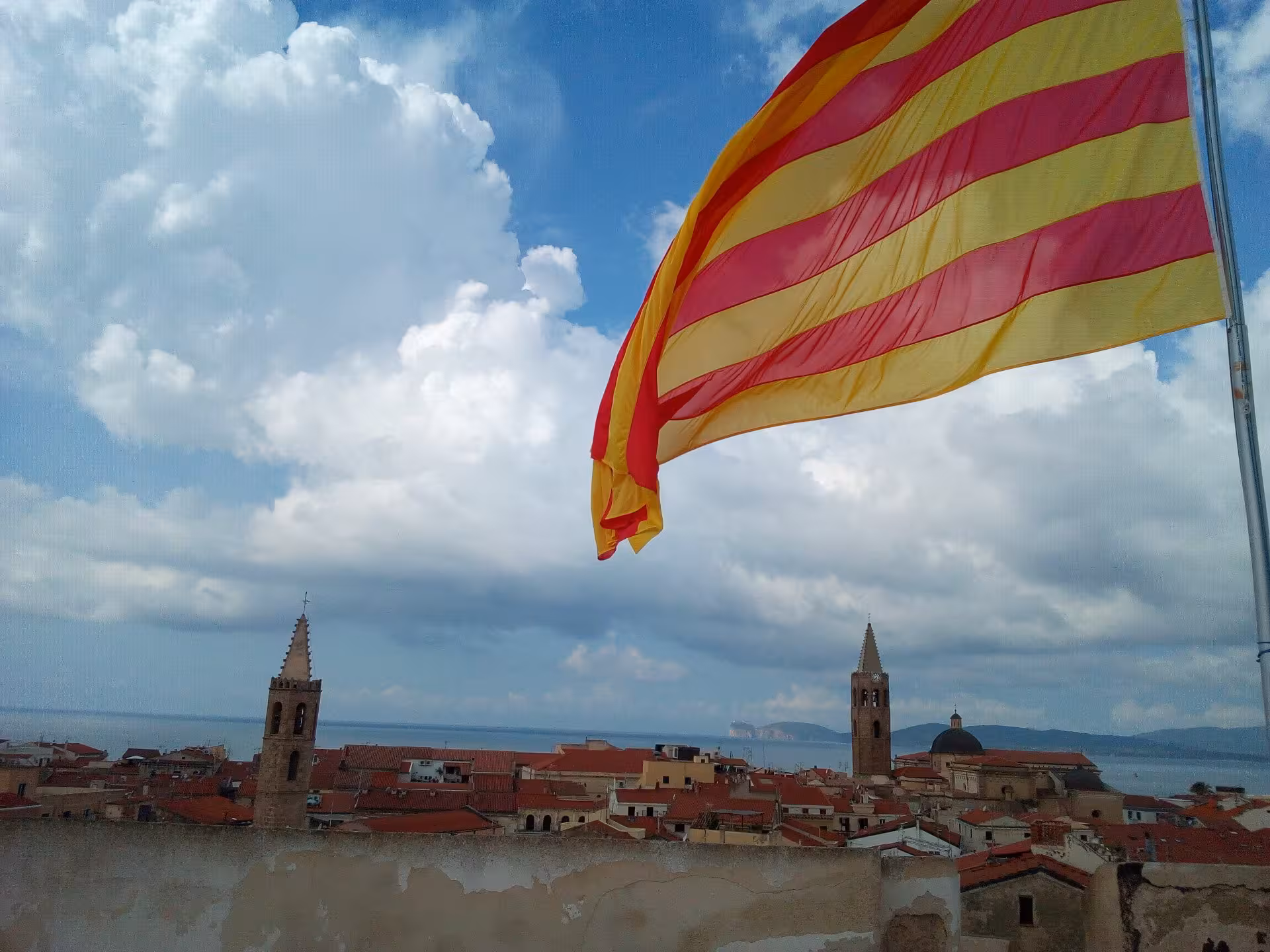 Experience the vibrant skyline of Alghero with views of historic towers and the iconic Catalan flag waving.