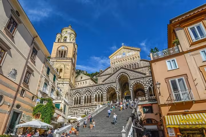 Majestic facade of Amalfi Cathedral with its grand staircase, surrounded by historic buildings under a vibrant blue sky.