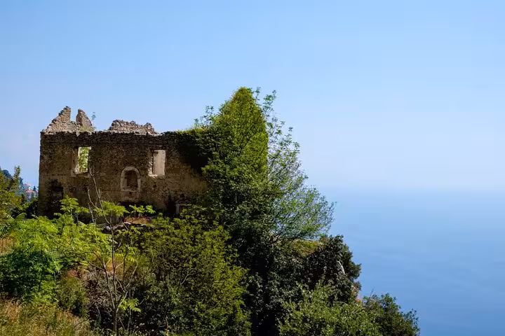 Ruins of a historic stone building surrounded by lush greenery overlooking the expansive blue sea on the Amalfi Coast.