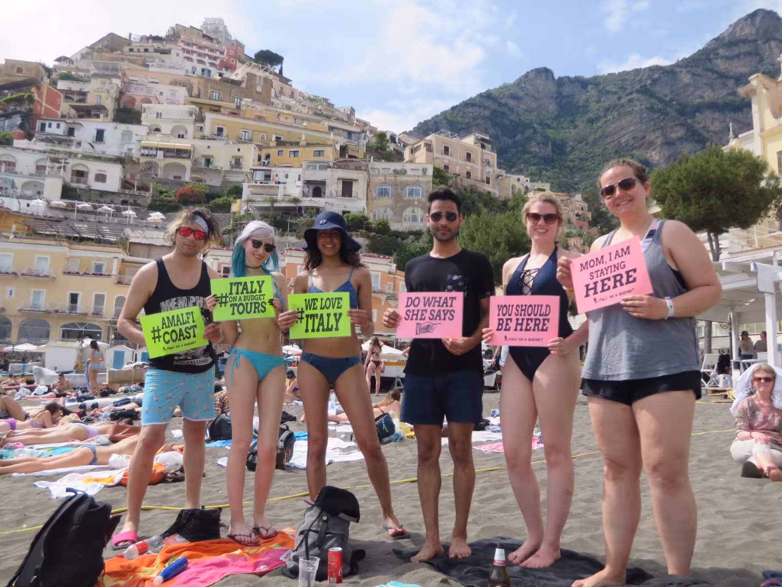 Tourists holding signs at Positano beach with colorful cliffside houses on the Amalfi Coast.