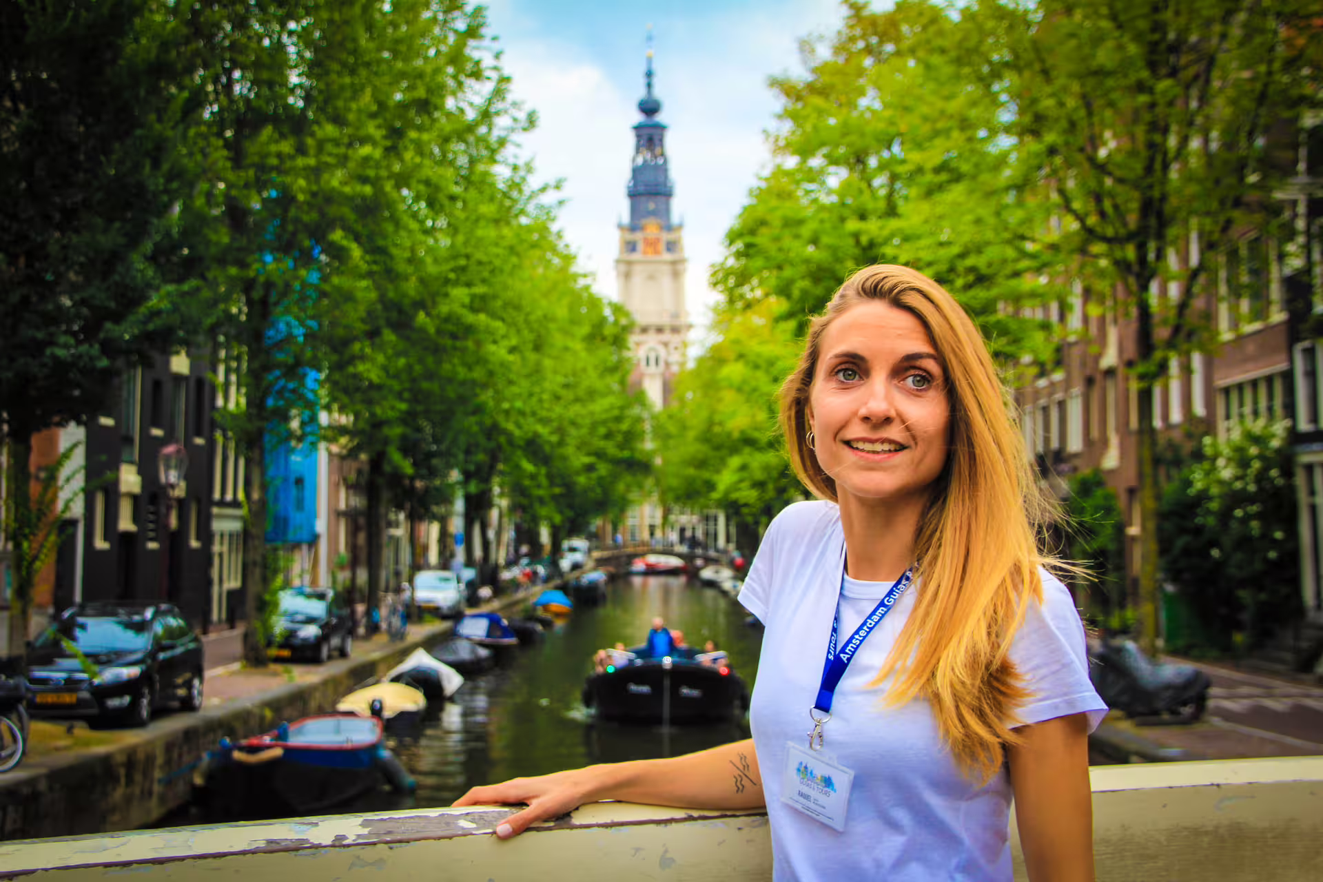 Smiling tour guide in Amsterdam with scenic canal, lush trees, and historic architecture, highlighting bike and canal cruise tour.