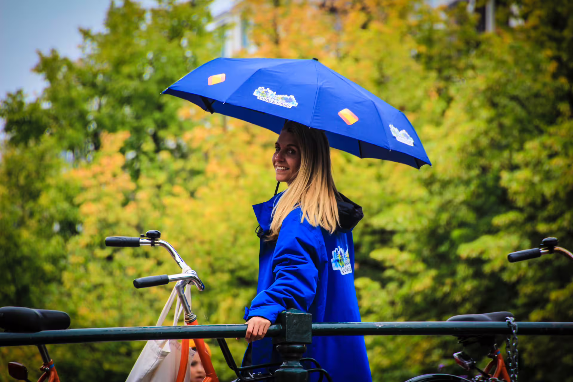 Smiling guide in blue coat and umbrella leads an Amsterdam bike tour with vibrant greenery in the background.
