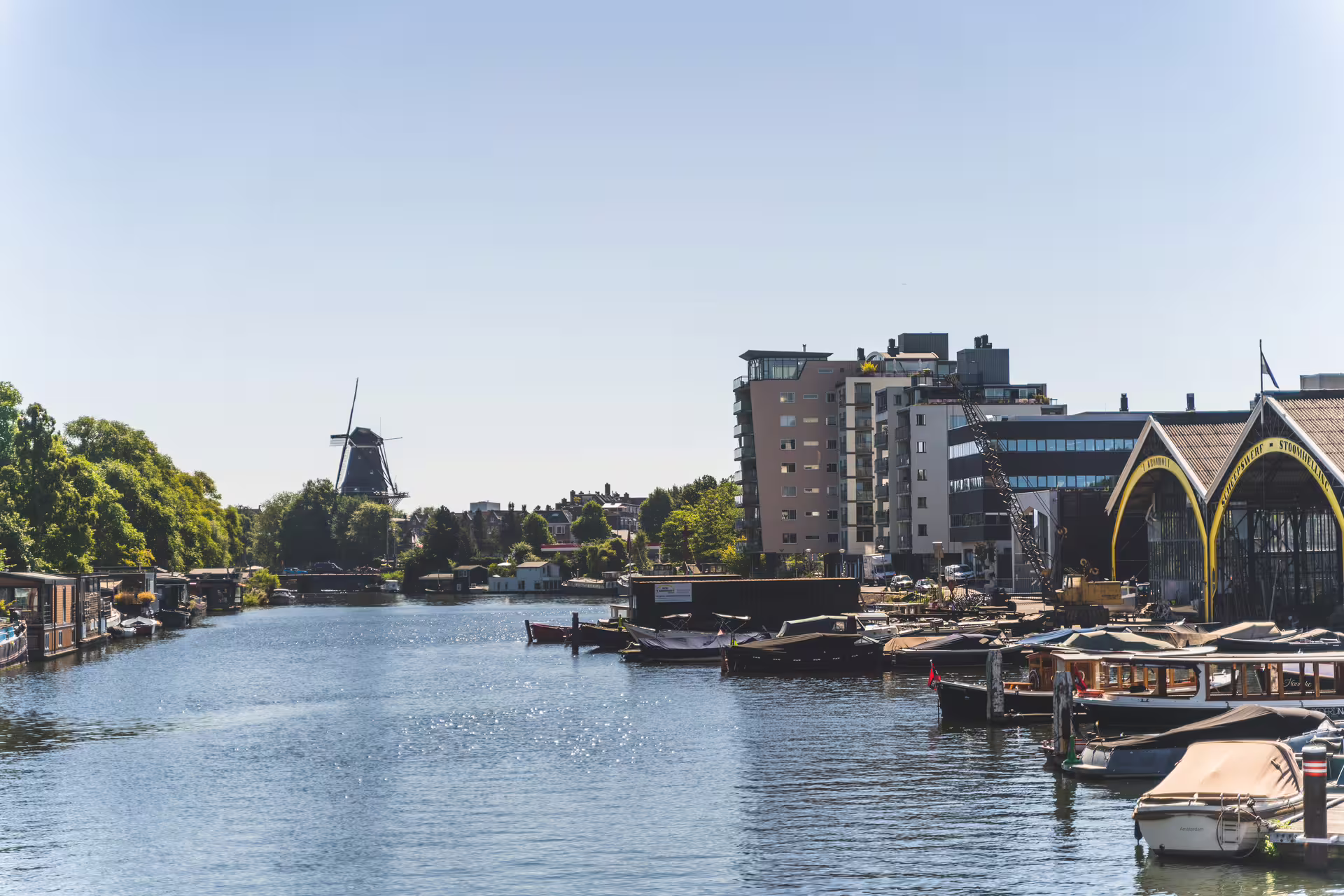 Scenic view of Amsterdam canal with boats, historic windmill, and modern buildings, perfect for bike tour and canal cruise experience.