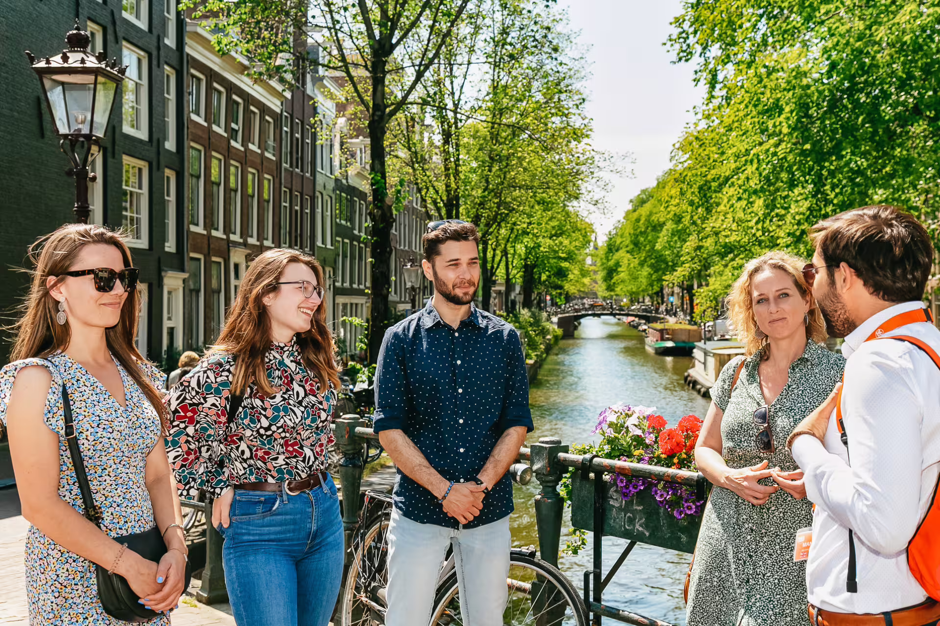 Tour group exploring historical Amsterdam, standing by a picturesque canal under sunny skies, led by a local guide.