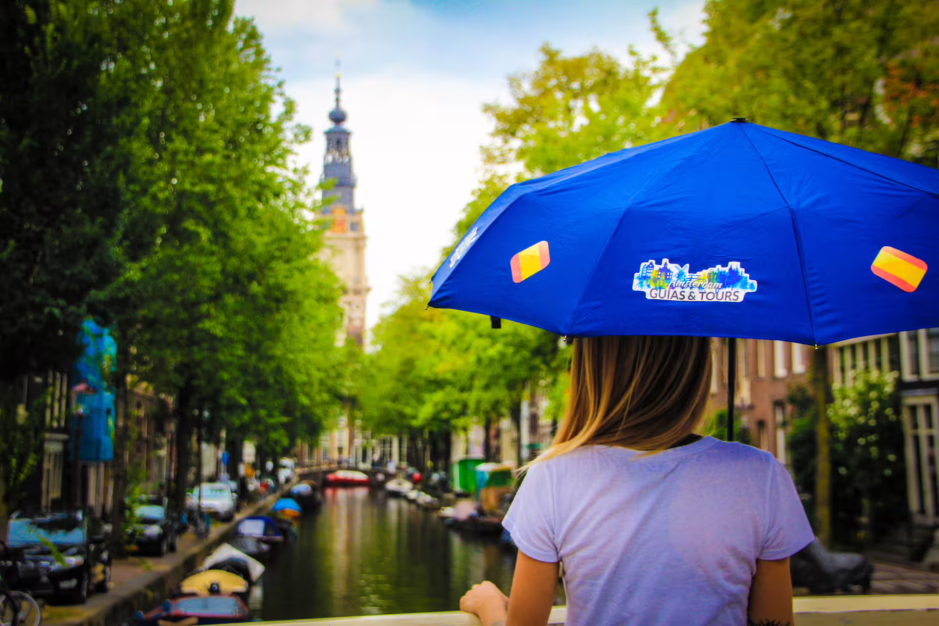 A person holding a blue umbrella with a tour logo overlooks a picturesque Amsterdam canal, highlighting a private walking tour experience.