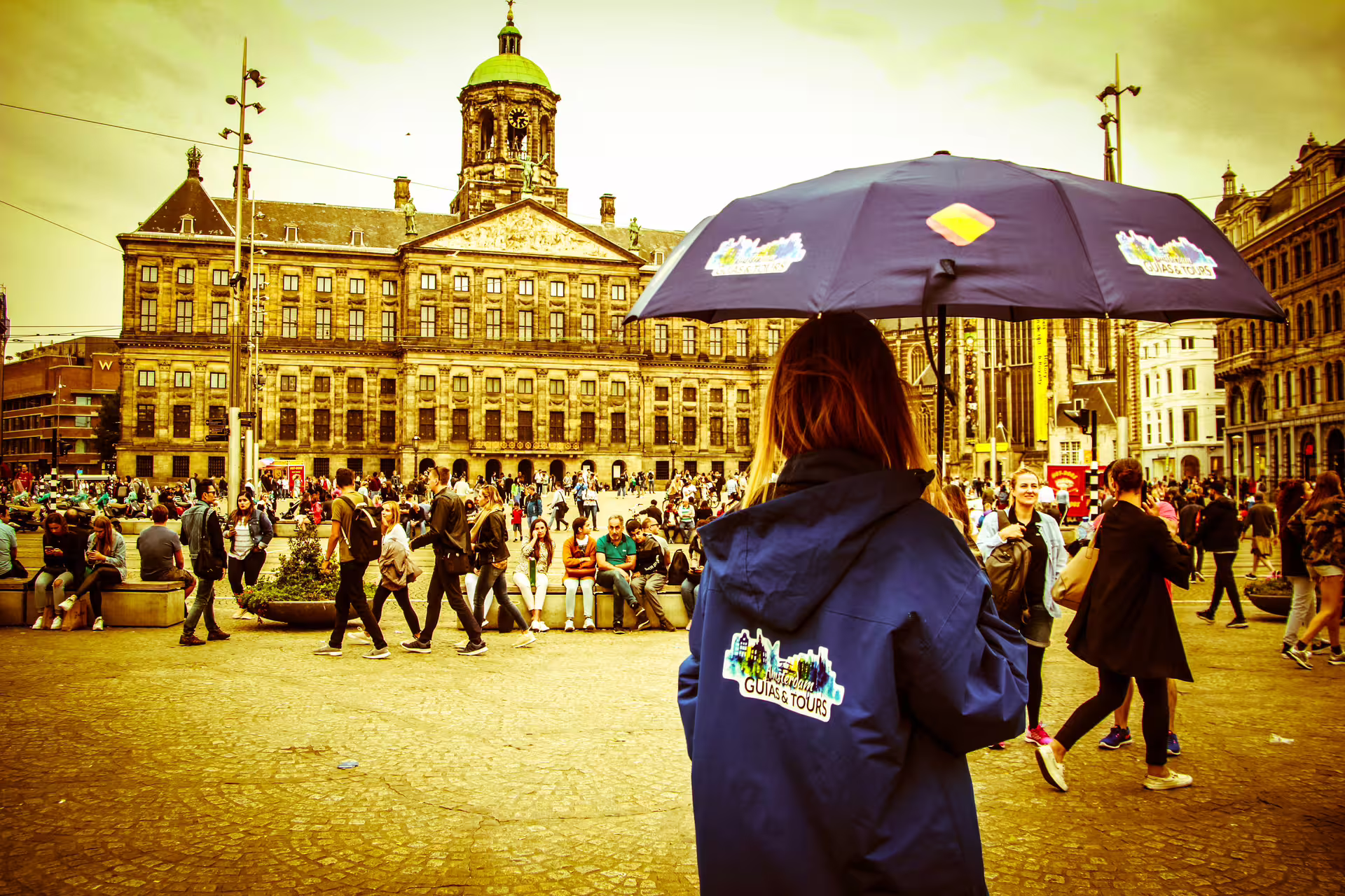 Tour guide in blue jacket leads private walking tour in Amsterdam's bustling Dam Square, showcasing historic architecture.
