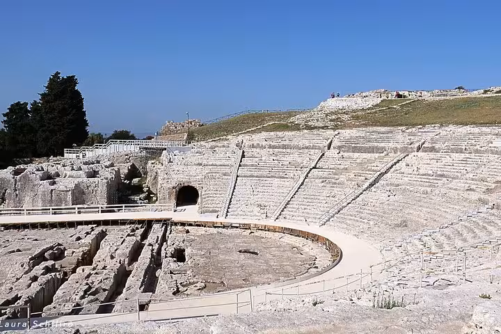 Ancient Greek theater in Syracuse under blue sky, showcasing stone seating and historical architecture.