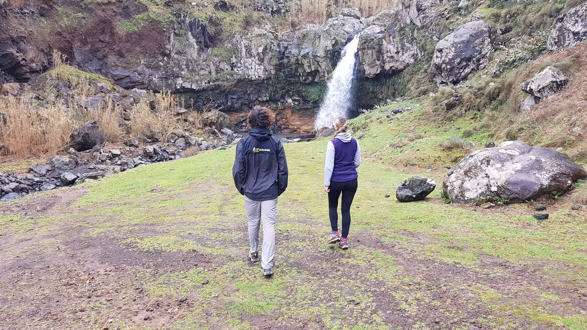 Two hikers approach a cliffside waterfall across an open green meadow on the HIKE Lomba de São Pedro - Moinhos Félix