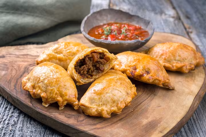 Golden Argentinian empanadas filled with savory meat, served with a spiced tomato dipping sauce on a rustic wooden board.
