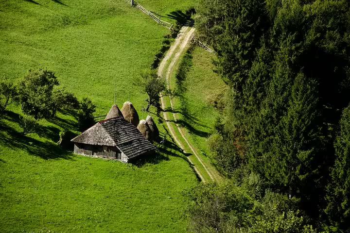Aerial view of a traditional Romanian wooden farmhouse with haystacks on a lush green hillside with a dirt road