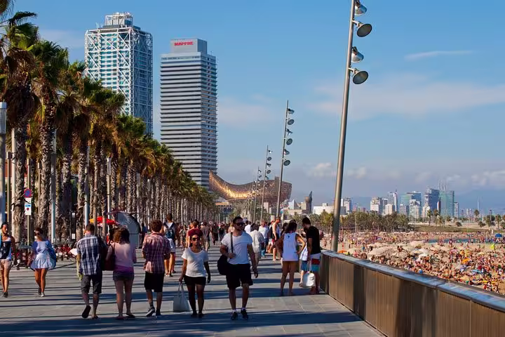Tourists walk along the bustling Barcelona beachfront promenade with skyscrapers and palm trees, illustrating urban development and leisure.