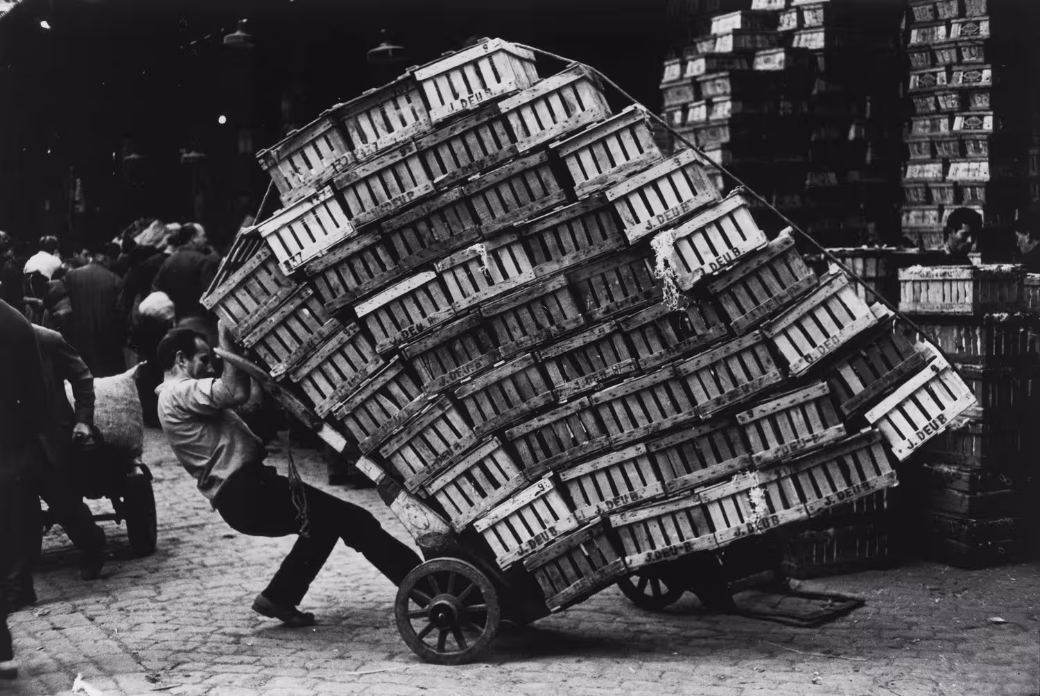 Vintage market scene in Barcelona's Ribera, highlighting a worker balancing a large stack of wooden crates on a cart.