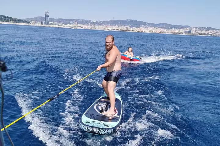 Man paddleboarding behind a boat on a sunny day in Barcelona, with city skyline and mountains in the background, enjoying water activities.