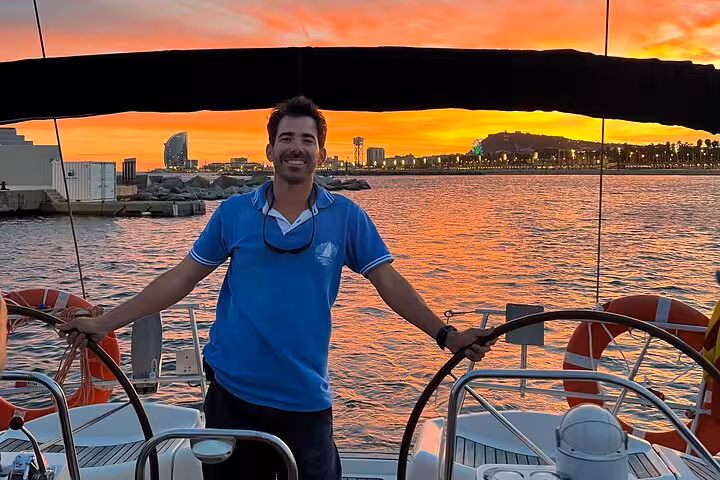 Skipper on Barcelona sunset sailing tour with open bar and snacks, city skyline glowing behind him