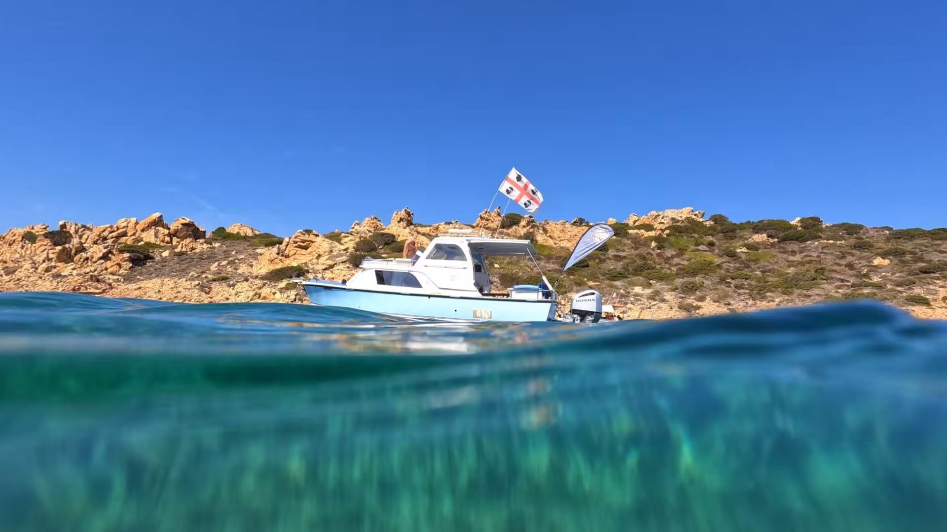 A blue speedboat anchored near rocky shores in the La Maddalena Archipelago under a clear blue sky.