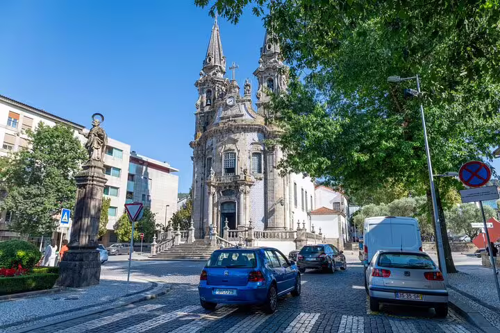 Baroque church with twin spires in Braga surrounded by lush greenery and parked cars.