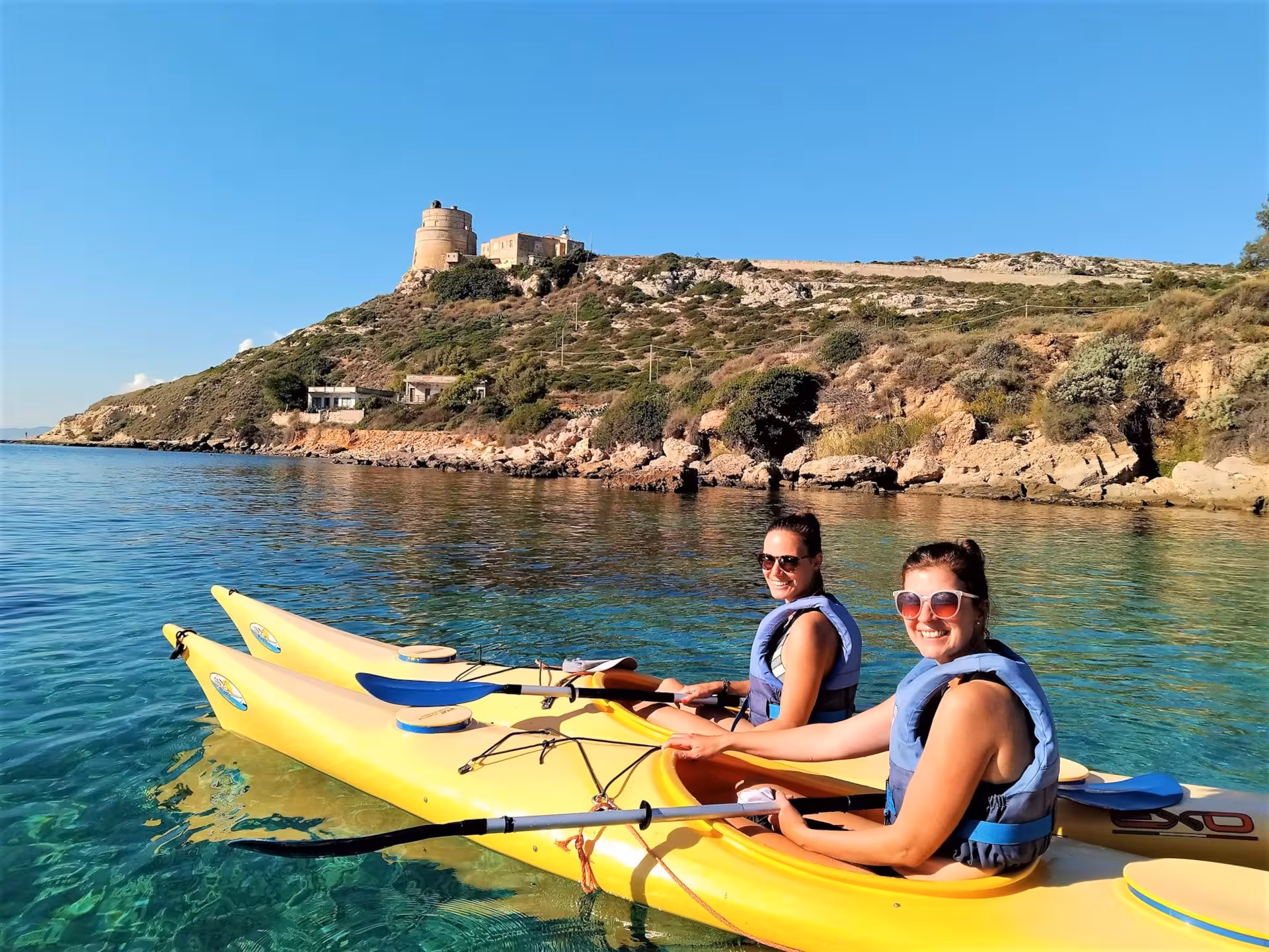 Two women in kayaks paddle near a historic tower on Cagliari's scenic coastline in sunny weather.