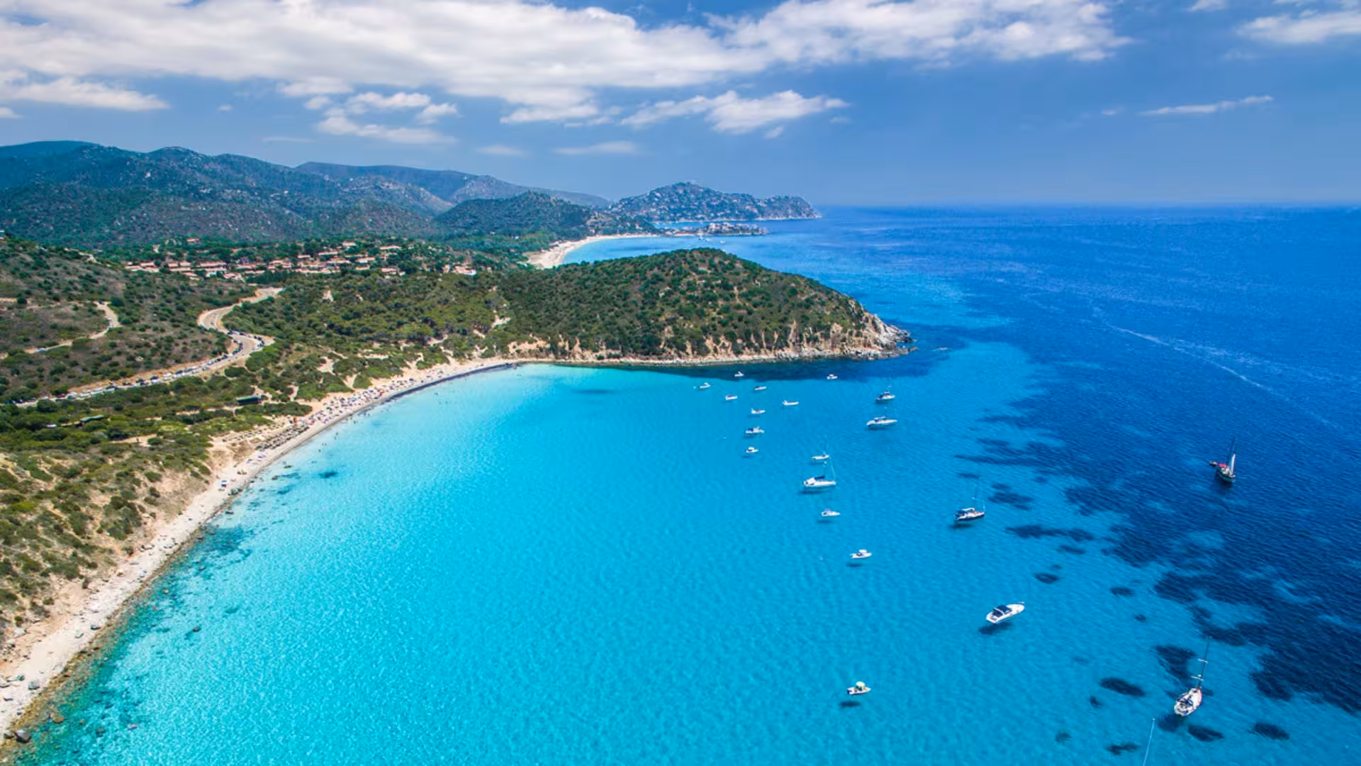 Aerial view of Mari Pintau's turquoise waters with sailing boats, lush coastline, and scenic hills in Cagliari.