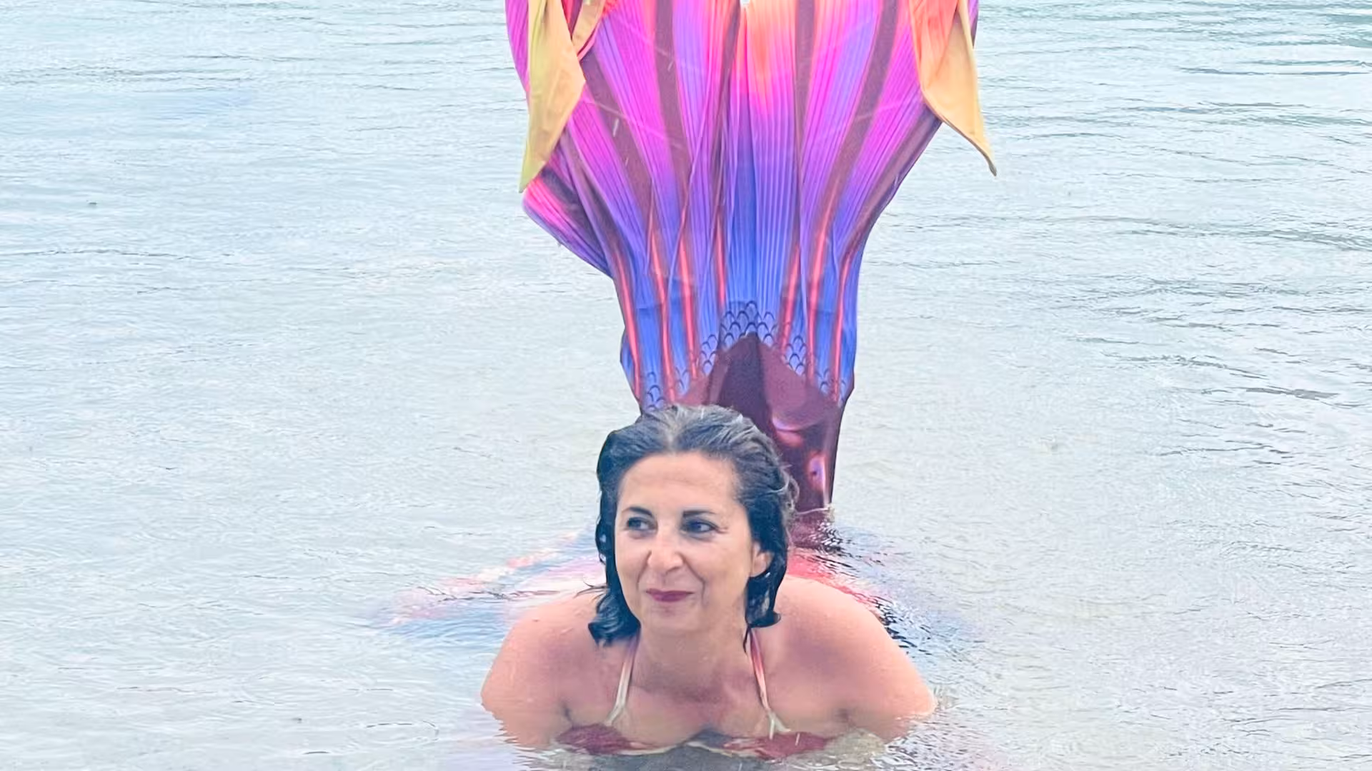 Woman with a vibrant mermaid tail relaxing in shallow waters during a Cagliari mermaid experience.