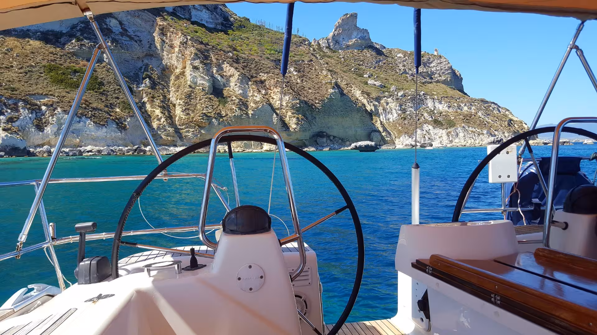View from a sailing boat navigating the azure waters near Mari Pintau, showcasing rugged cliffs and clear skies.
