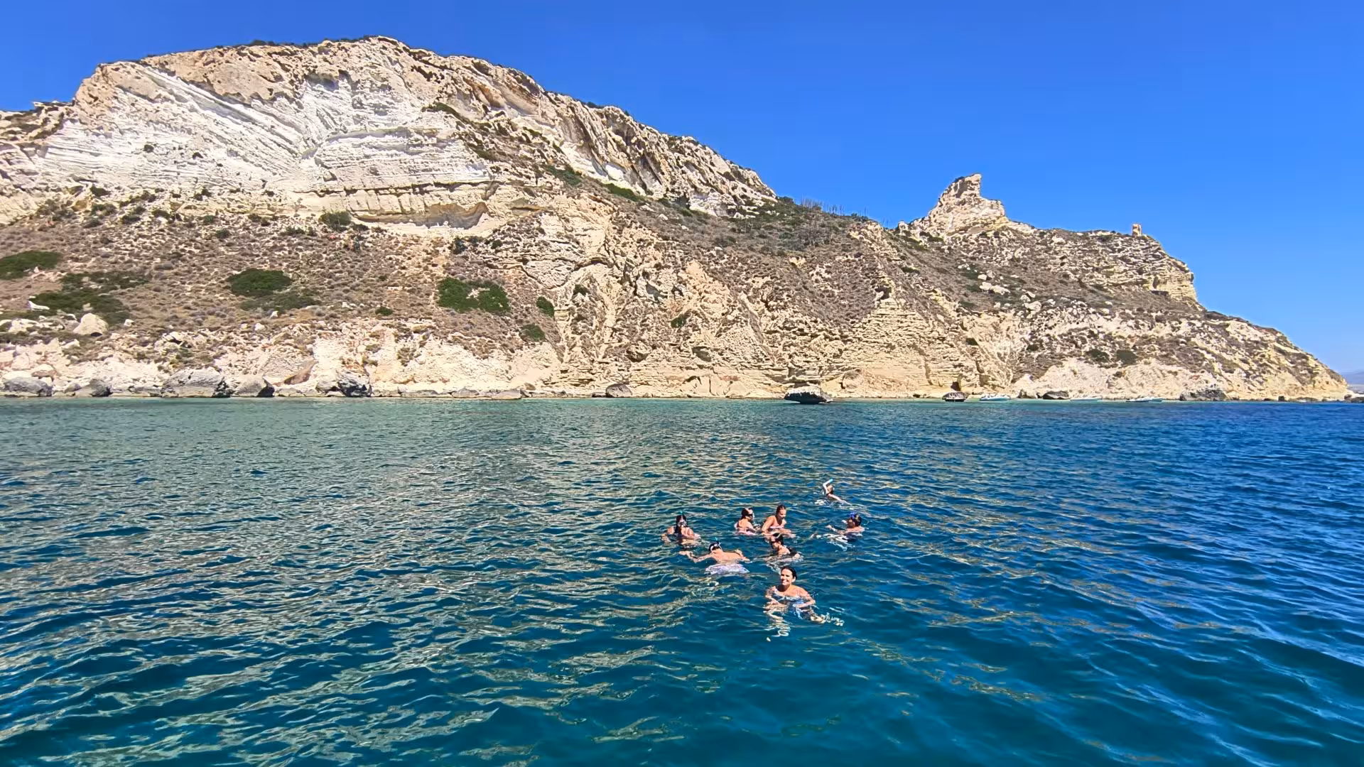 Group of swimmers exploring the vibrant blue waters against the scenic cliffs of Cagliari on a sailing tour.