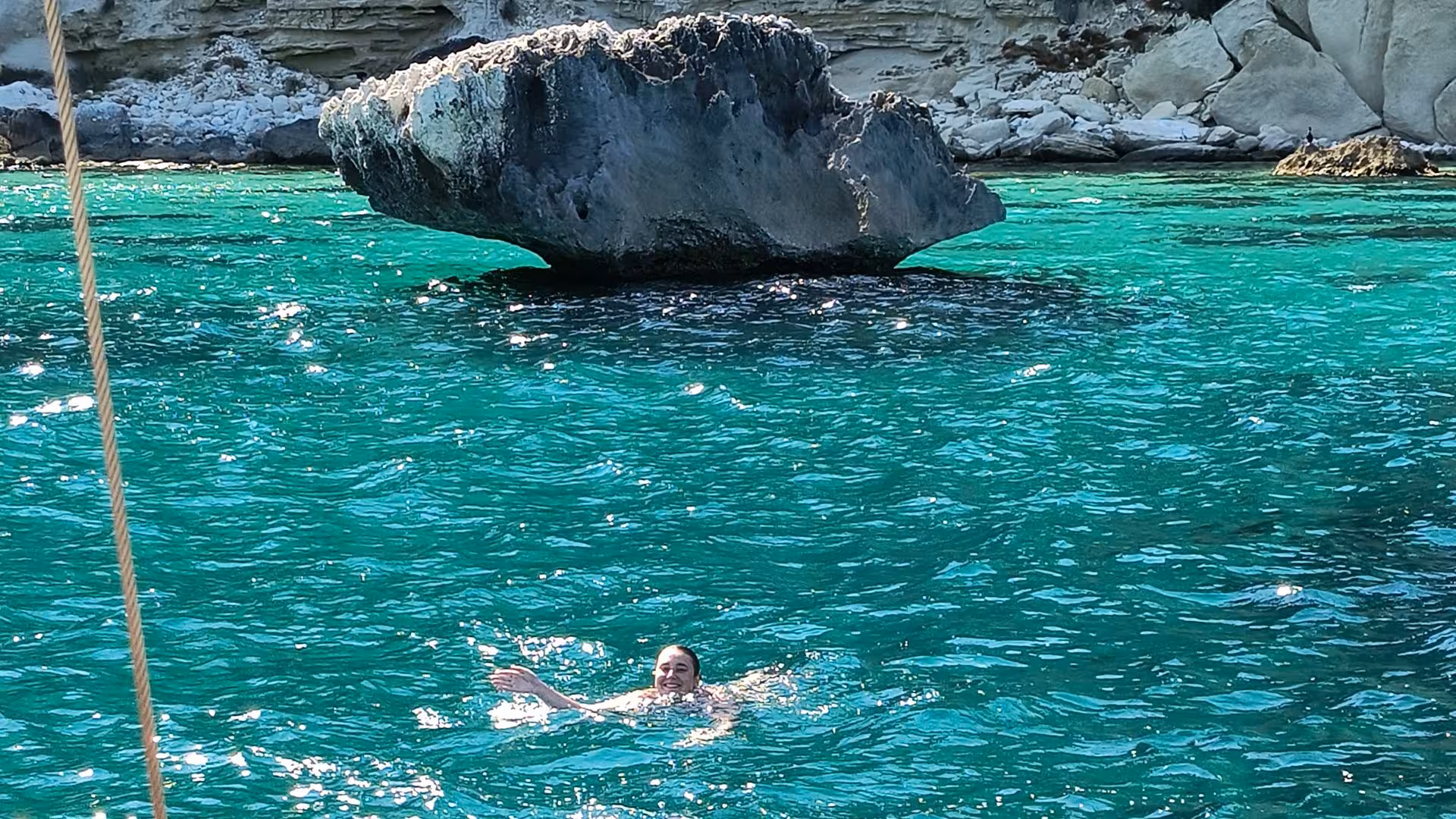 Swimmer enjoying crystal-clear turquoise waters near rocky Cagliari coastline during a sailing excursion.