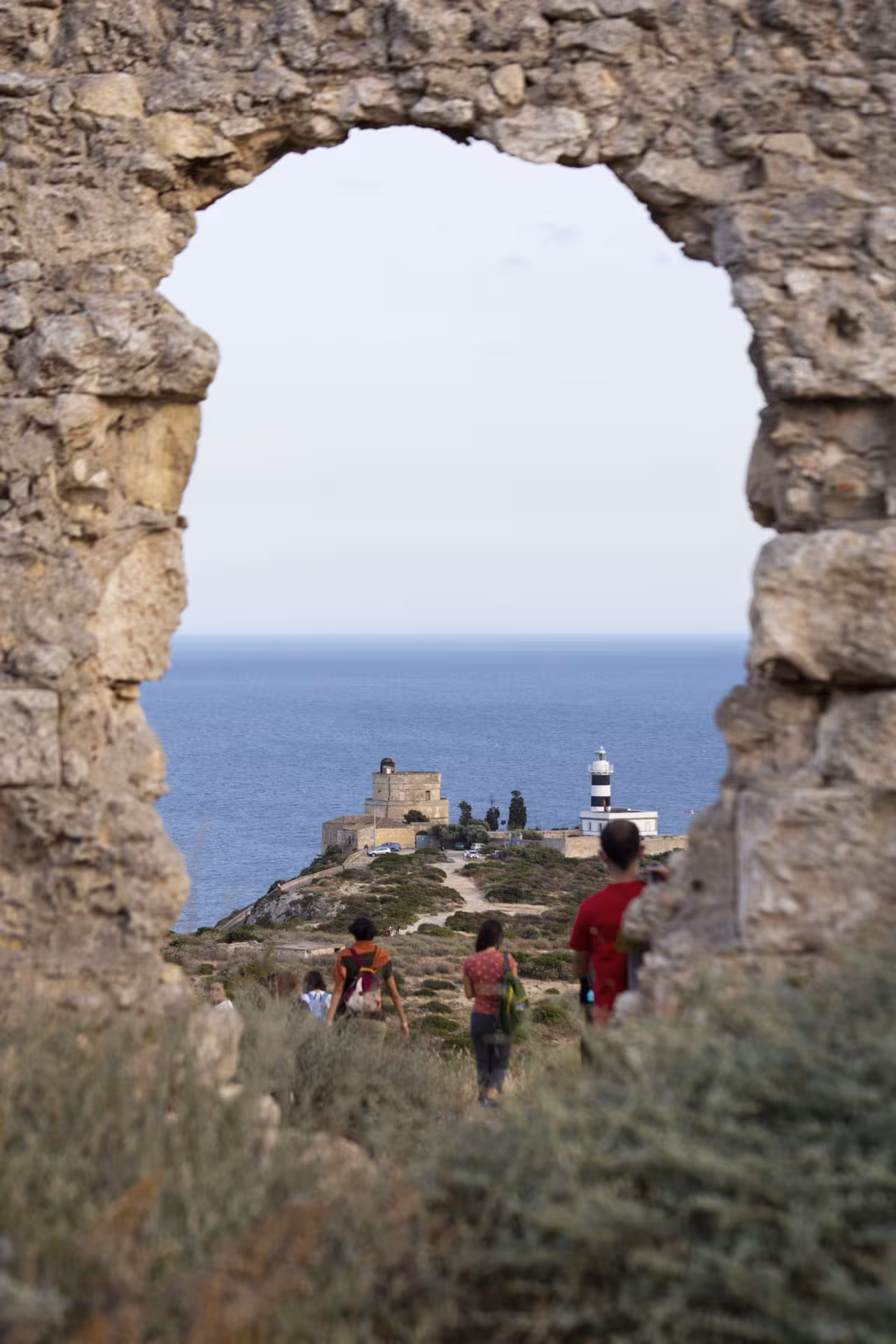View through ancient stone arch of Sant'Elia hill trek with hikers and lighthouse overlooking Cagliari's coastline.
