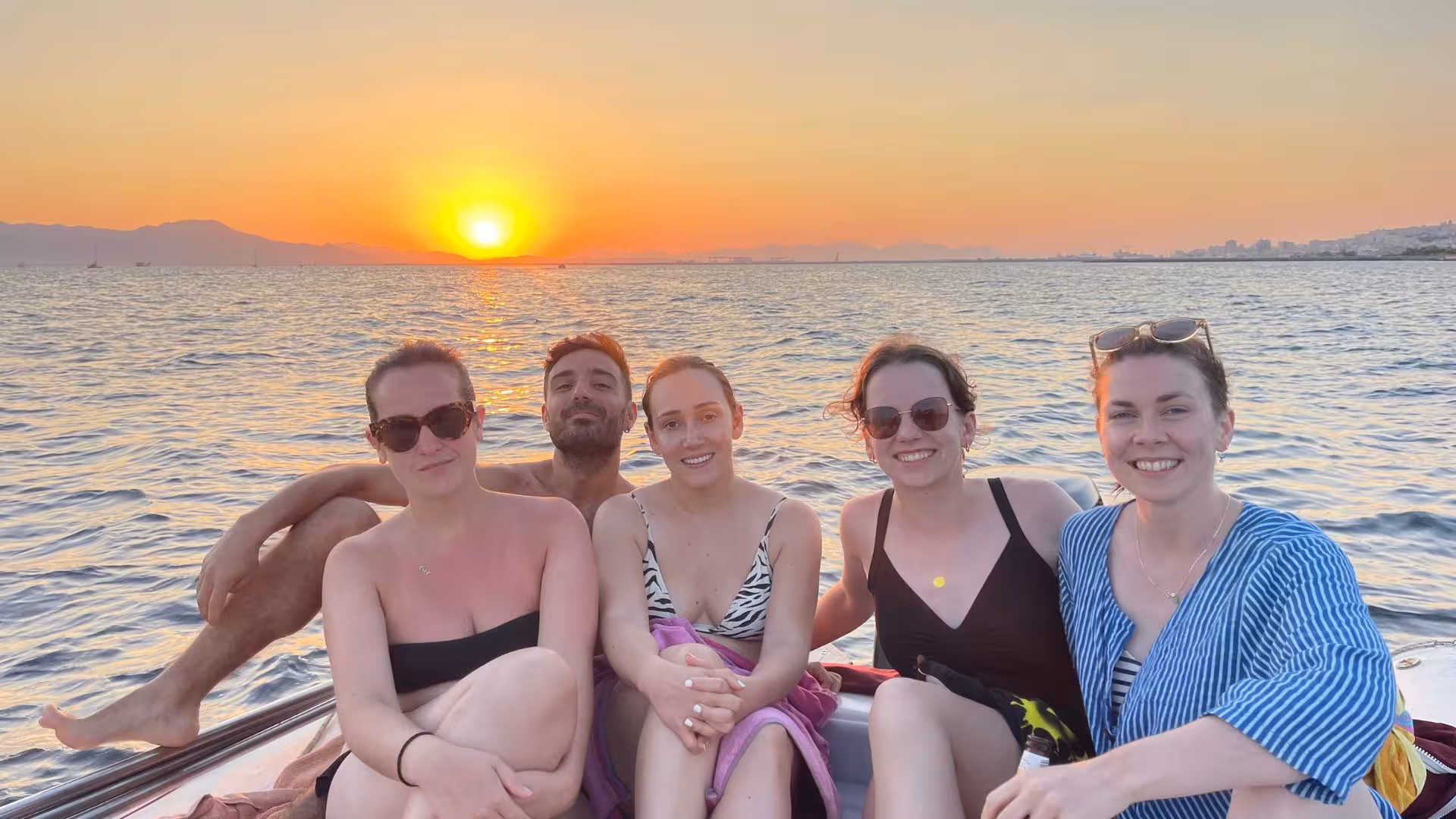 Happy group on a dinghy at sunset near Cagliari, with scenic views and a warm glow reflecting on the sea.