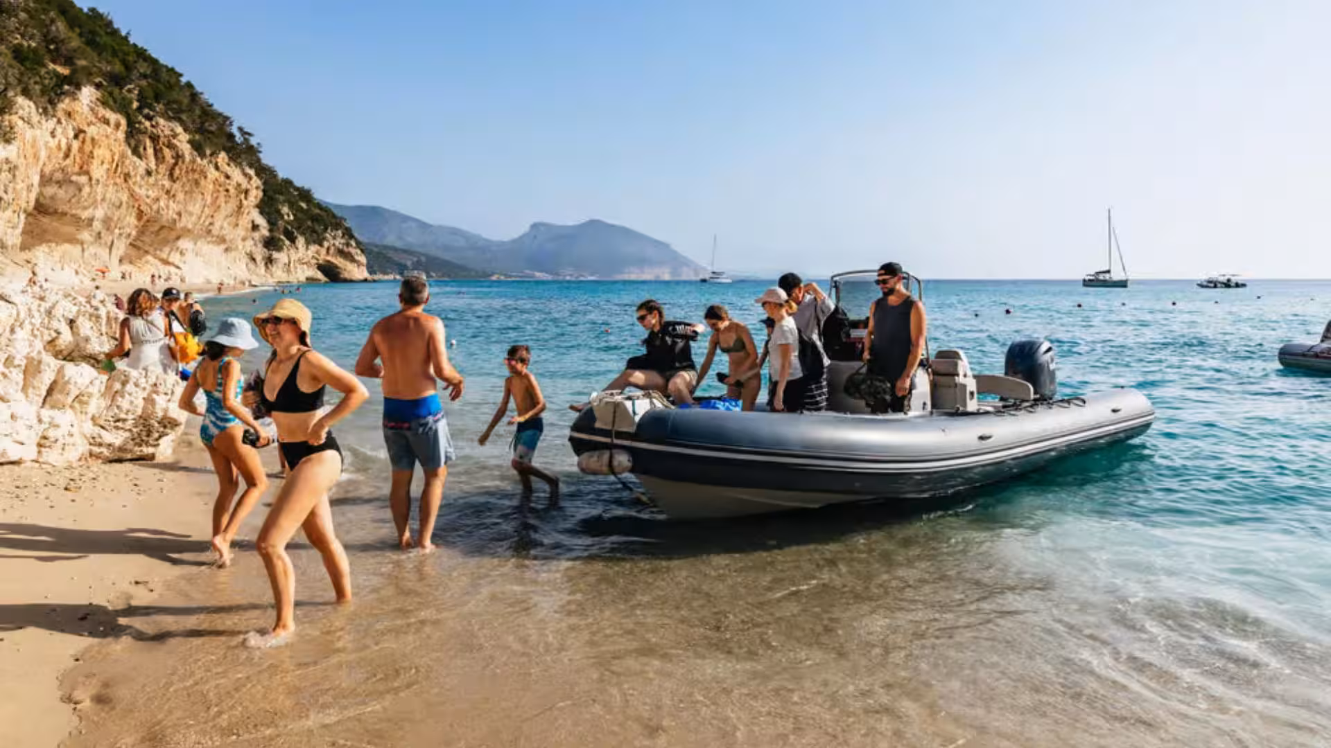 Tourists disembarking from a dinghy onto a sandy beach in Cala Gonone, surrounded by scenic cliffs.