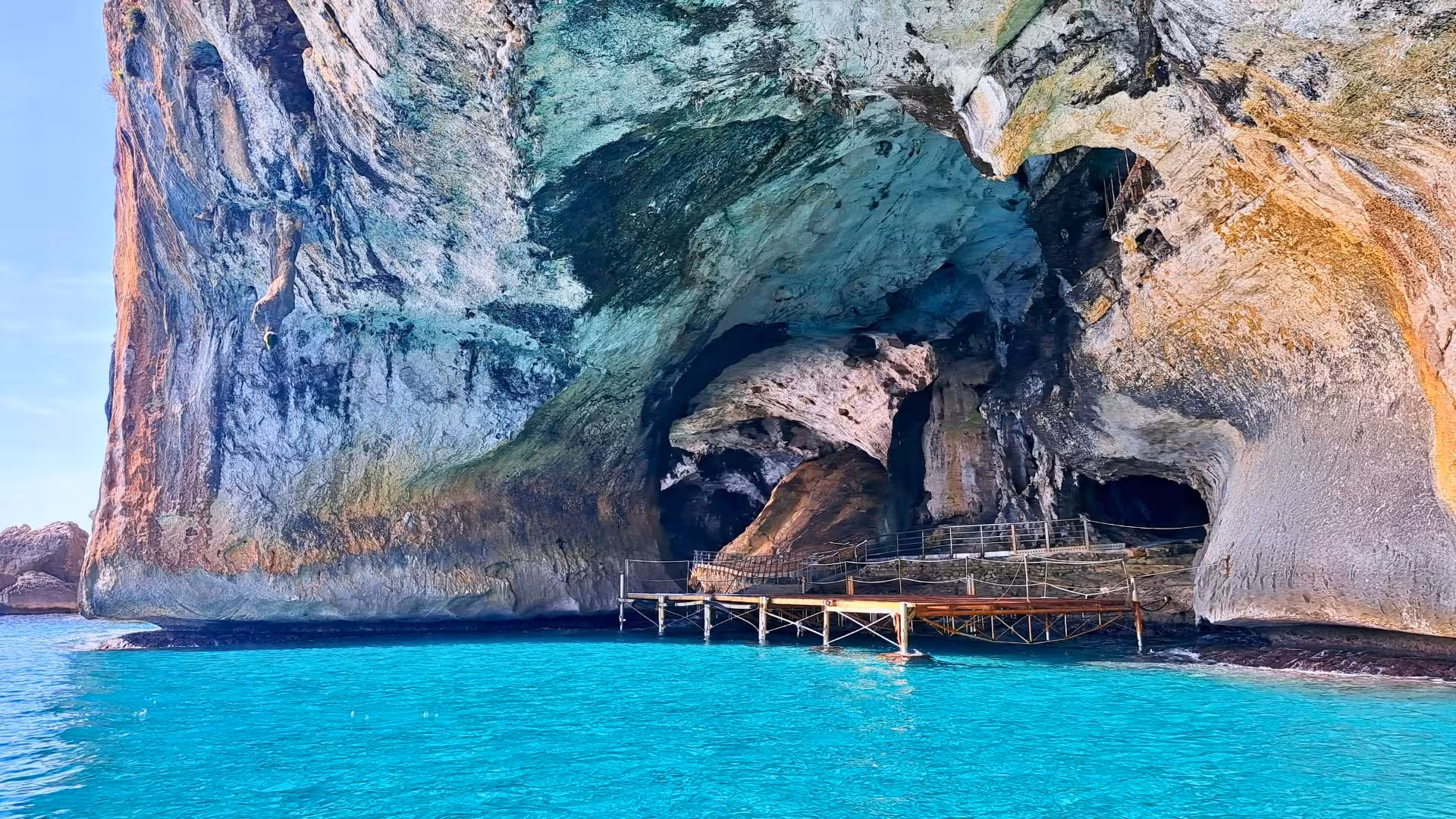 Scenic view of a wooden pier leading into a large rock cave with vibrant turquoise waters in the Gulf of Orosei.