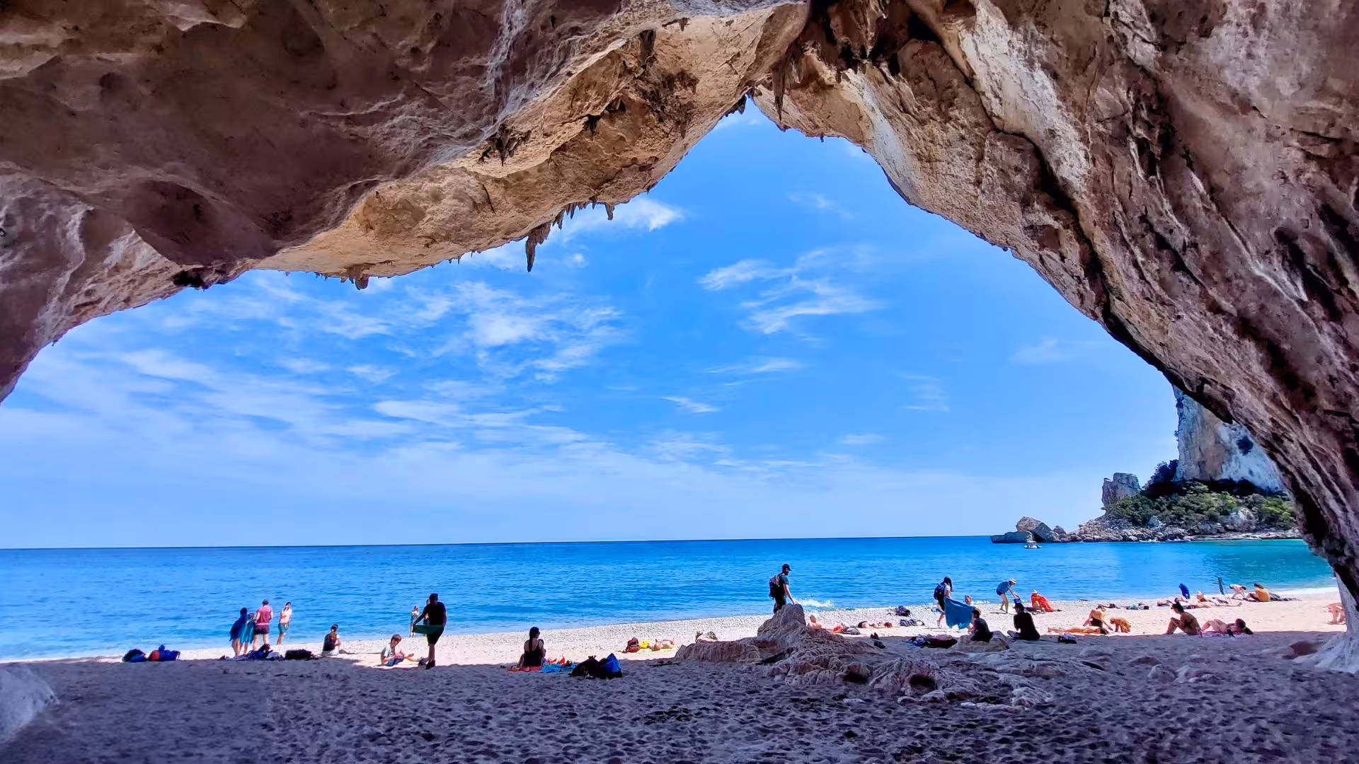 Visitors relax on a sandy beach under a natural rock arch, overlooking the blue waters of the Gulf of Orosei.